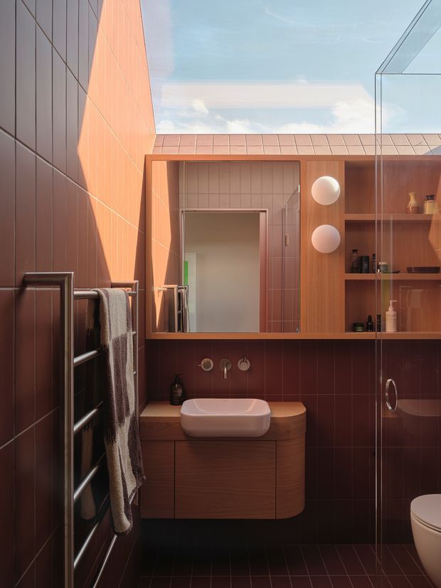 Compact contemporary bathroom with warm terracotta wall and floor tiles, a timber floating vanity with a white vessel basin, wall-mounted fixtures, open timber shelving, and a glass shower enclosure. A skylight or glazed roof opening brings in daylight, while a heated towel rail and partially visible toilet complete the space.