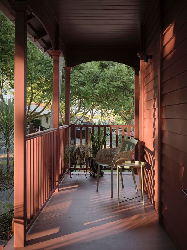 A covered timber porch or veranda viewed along its length, with reddish-brown weatherboard siding, matching timber railings and posts, an arched opening, and a dark timber-lined ceiling. The porch contains two woven lounge chairs, a small round table, and potted plants. Warm late-day sunlight creates strong shadows across the floor and walls, with leafy garden surroundings visible beyond the railing.