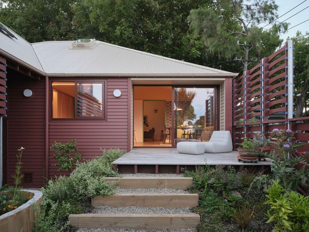 A compact single-storey residential pavilion viewed from a landscaped garden. The exterior has dark reddish-brown horizontal weatherboard cladding, a light grey corrugated metal pitched roof, and large glazed openings connecting to a raised timber deck. The deck includes light upholstered outdoor seating, while timber privacy screening with horizontal slats encloses one side. Stepping-stone and timber-edged stairs lead through dense planting toward the house. Warm interior lighting is visible through the open glazed doors and windows.