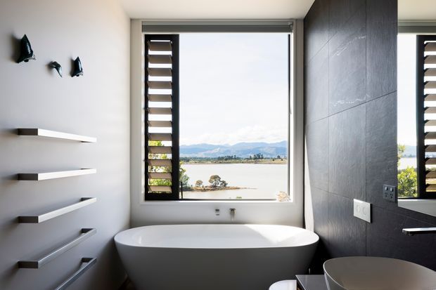 A contemporary bathroom with a freestanding white bathtub positioned beneath a large picture window. Dark tiled walls contrast with white walls and floating shelves, while a louvred window panel and mirror add architectural detail. The window overlooks a broad body of water and distant mountains.