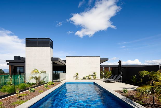 A contemporary single-storey residence viewed from the outdoor pool area. The image features a long rectangular swimming pool, pale rendered or concrete geometric wall volumes, dark horizontal cladding and roof elements, extensive glazing, paved pool surrounds, and sparse tropical-style landscaping under a bright blue sky.