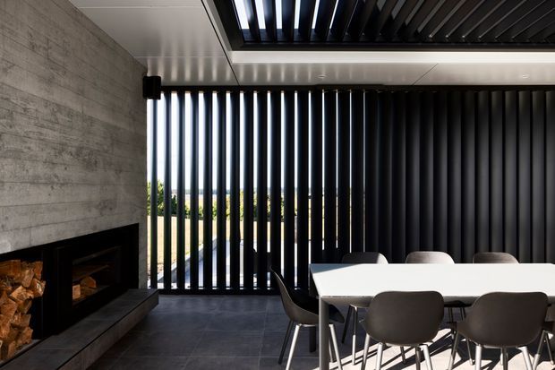 A contemporary dining area beside a dramatic built-in fireplace, featuring a long white dining table with dark upholstered chairs. The room has a dark tiled floor, a raw concrete feature wall, black vertical louvres screening the glazing, and a black slatted ceiling element. Daylight and greenery are visible beyond the louvres.
