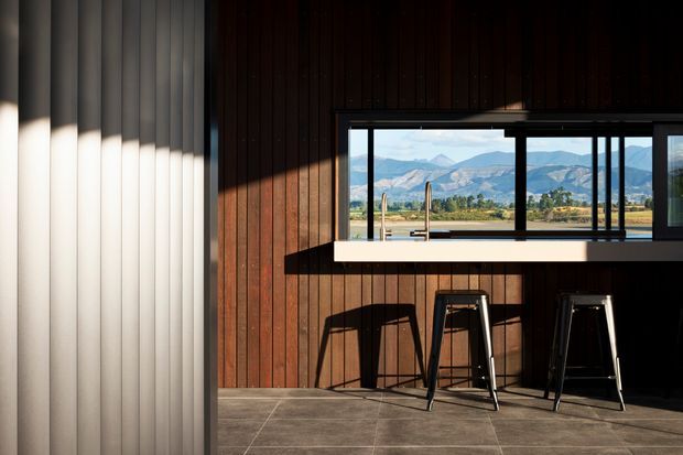 A contemporary interior bar or kitchen counter area with dark vertical timber wall panelling, a white benchtop, black-framed horizontal glazing, and two black stools. The window provides a clear view toward a mountainous landscape and open rural surroundings. Strong sunlight creates dramatic shadows across the walls and tiled floor.