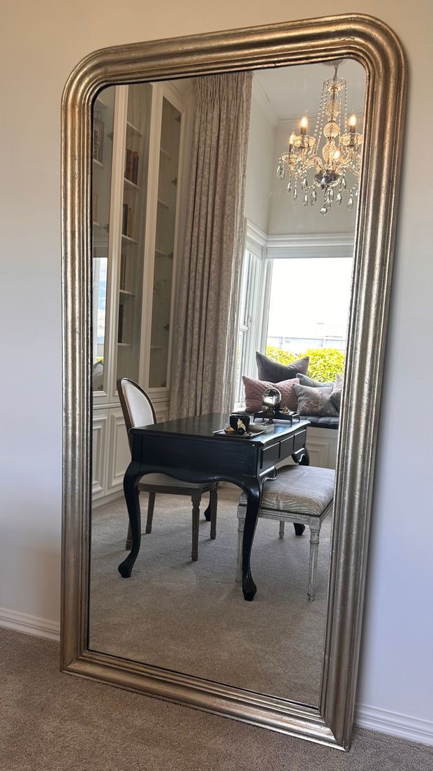 A tall ornate framed mirror reflects an elegant, traditional interior featuring a black writing desk with curved legs, upholstered chairs, built-in shelving, full-length patterned curtains, a window seat with cushions, and a crystal chandelier. The visible room has soft neutral finishes and a refined, layered decorative style.