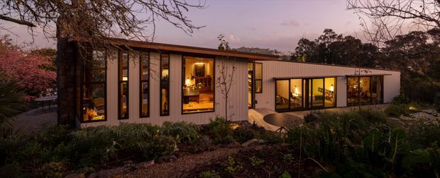 A low, elongated contemporary house nestled into a landscaped garden at dusk. The facade combines dark vertical cladding, pale panelled walls, extensive floor-to-ceiling glazing, and a shallow low-pitched roof. Warm interior lighting glows through the windows, revealing living spaces and built-in joinery, while a curved paved path leads toward the entrance.