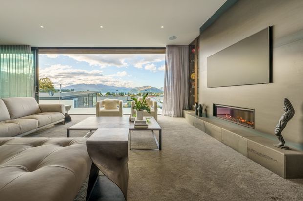 A refined contemporary living room with a large sectional sofa and ottoman, low coffee table, wall-mounted television, and built-in linear fireplace. Full-height glazing and a balcony open to expansive mountain and water views, while sheer curtains, recessed ceiling lights, neutral upholstery, and stone-toned finishes create a calm, luxurious atmosphere.