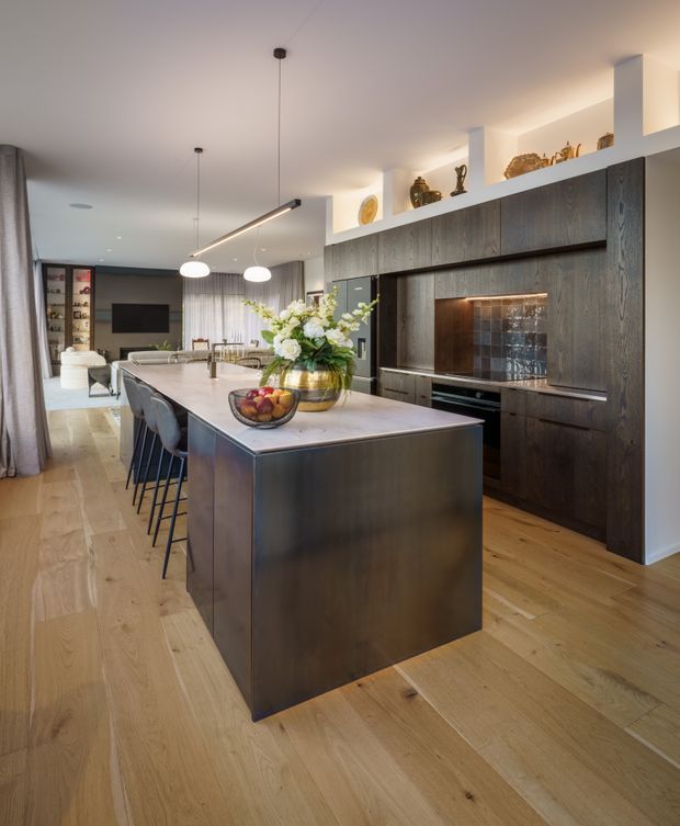 Contemporary open-plan kitchen with a large dark timber island, integrated cabinetry, pale stone benchtop, dark tiled splashback, and light timber flooring. The kitchen opens into a dining and living area, with pendant lighting, white structural columns, display shelving, and restrained decorative styling.