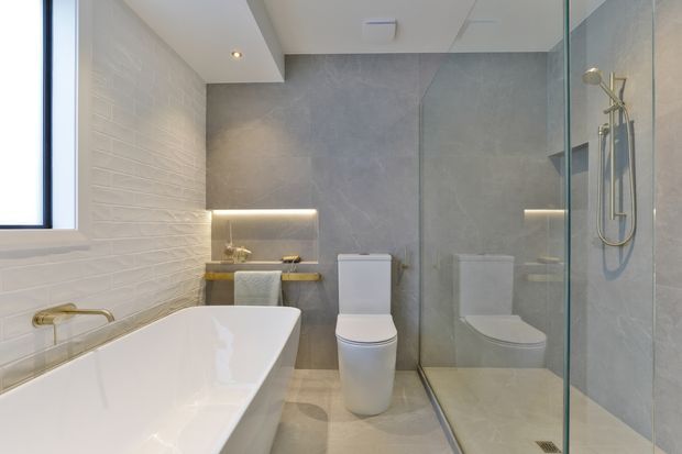 Bright contemporary bathroom featuring a freestanding white bathtub, wall-mounted brass tapware, a wall-hung toilet, large walk-in glass shower, and illuminated recessed shelving. The room uses light grey stone-look wall and floor finishes, white textured wall tile, and brushed brass fixtures.