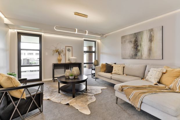 Bright contemporary living room with a large light-grey sectional sofa, black round coffee table, sculptural lounge chairs, layered neutral textiles, abstract wall art, and a slim linear pendant light. Floor-to-ceiling windows and concealed perimeter lighting create an airy, refined atmosphere, with a dark slatted console and warm beige accents adding contrast.