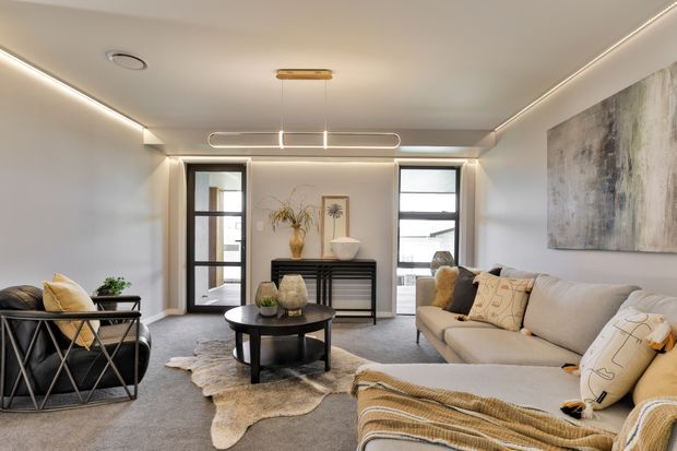 A contemporary living room with a low, softly lit ceiling, recessed perimeter lighting, dark-framed glazed doors and windows, a black console, round coffee table, layered neutral seating, and a large abstract artwork. The room has a calm, refined neutral palette with grey carpet, cream upholstery, black accents, and warm metallic lighting details.