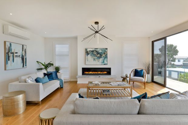Bright contemporary living room with pale upholstered sofas, blue accent cushions, a timber floor, a linear electric fireplace set into a white feature wall, abstract artwork, plantation shutters, and large black-framed sliding glass doors opening to an outdoor view. A sculptural starburst pendant light forms a central focal point.