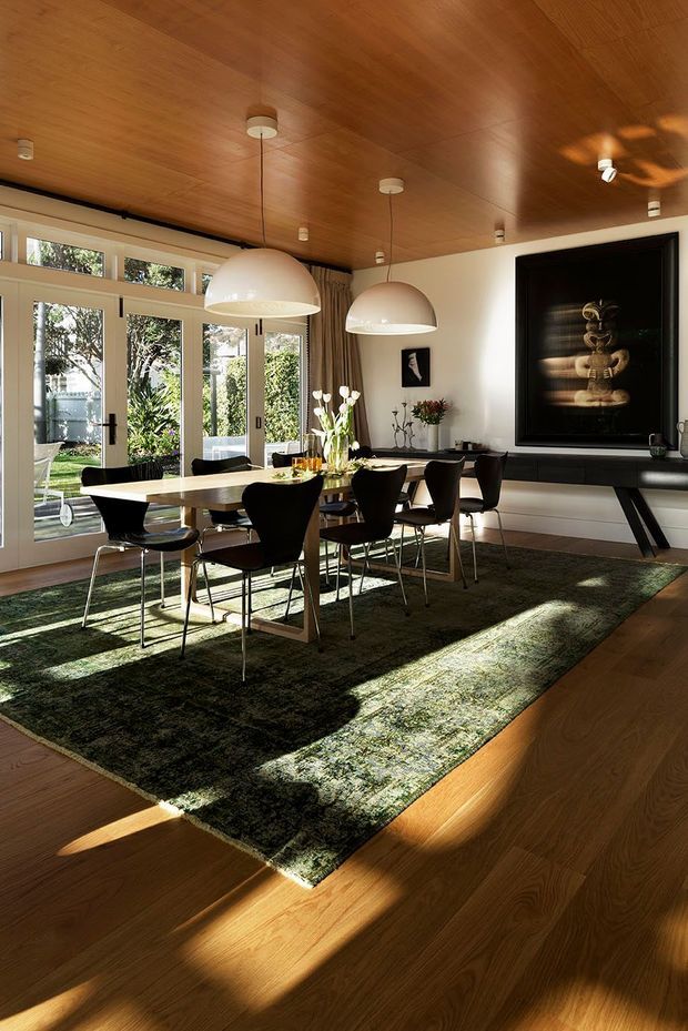 A bright, refined dining room with a long light-toned dining table, sculptural dark dining chairs, oversized white pendant lights, and a large dark green patterned rug. Full-height glazed doors and windows open to a leafy garden, while a warm timber-lined ceiling and timber flooring create a cohesive natural palette. A large framed artwork and sideboard occupy the far wall.