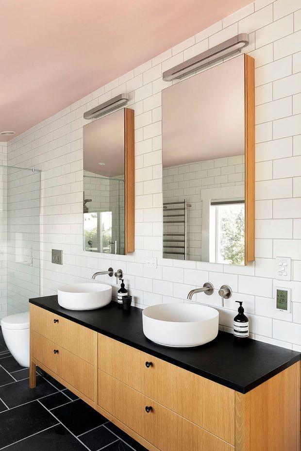 Bright contemporary bathroom featuring a double vanity with white vessel basins, black benchtop, timber cabinetry, white subway-tiled walls, black floor tiles, wall-mounted mirrors and chrome fixtures. A glass shower enclosure, toilet and heated towel rail are also visible.