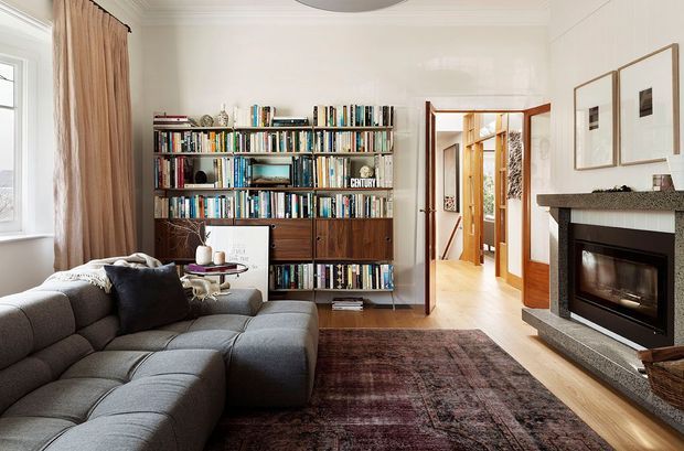 A warm, traditional living room with a large charcoal-grey sectional sofa, dark timber built-in bookshelves filled with books, a patterned area rug, and a stone-framed fireplace. White walls, timber flooring, framed artwork, and full-height curtains create a refined, lived-in interior. An open doorway leads to a hallway and additional rooms.