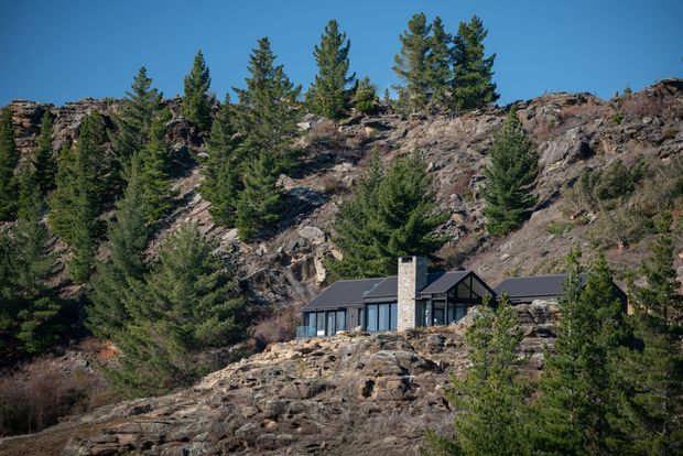 A modern dark-roofed house with extensive glazing and a prominent stone chimney is nestled into a rugged rocky mountainside among dense evergreen trees. The low-profile building blends into the alpine terrain, with exposed rock and forest dominating the surrounding landscape under a clear blue sky.