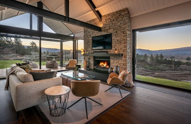 A spacious contemporary living room with expansive floor-to-ceiling glazing overlooking a rugged natural landscape. The room features a dramatic stacked-stone fireplace wall with integrated television and hearth, dark timber flooring, a pitched timber-lined ceiling, exposed dark structural beams, and a sophisticated seating arrangement with a large sectional sofa, leather lounge chairs, coffee table, and side tables. Warm firelight and sunset views create a refined, inviting atmosphere.