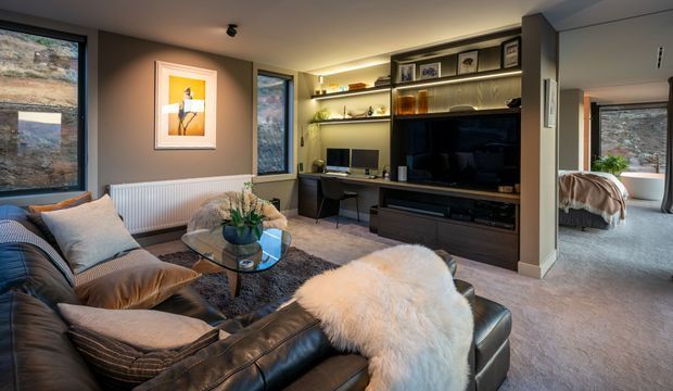 A warm, contemporary living room with a large dark leather sectional sofa, layered neutral cushions, a glass coffee table, and a sheepskin throw. Built-in dark timber entertainment joinery spans the rear wall, incorporating a television, open display shelving, study desk, books and decor. Large windows provide views toward a wooded landscape. A bedroom is visible through the opening on the right.