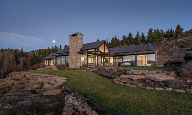 Exterior dusk view of a contemporary single-storey house integrated into a rocky, forested sloping site. The home has extensive floor-to-ceiling glazing, dark standing-seam pitched roofs, a prominent natural stone chimney, and a mix of stone and dark-framed glass walls. Layered stone landscaping and lawn surround the building.