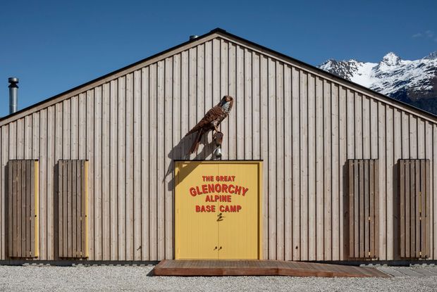 Front elevation of a simple gable-roofed alpine base-camp building clad in vertical pale timber boards. Multiple dark timber doors flank a central yellow entrance door with red lettering reading “The Great Glenorchy Alpine Base Camp,” beneath a mounted bird sculpture. Snow-capped mountains are visible behind the building under a clear blue sky.