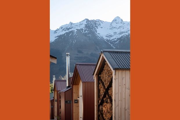 A vertically framed exterior view of a compact cluster of timber-clad buildings with steep pitched metal roofs, chimney flues, and a prominent open firewood storage wall. Snow-capped alpine mountains rise directly behind the buildings. The image has strong warm timber tones contrasted with cool blue-grey mountains and roofing, with orange side borders.