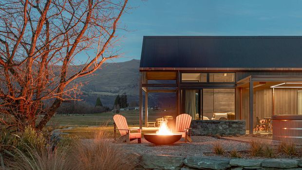 A contemporary single-storey mountain retreat with a dark metal gabled roof, extensive timber-framed glazing, and stone-clad exterior walls. The home opens onto a gravel terrace with an outdoor fire bowl, timber Adirondack chairs, and native-style landscaping, with rugged mountains visible beyond.