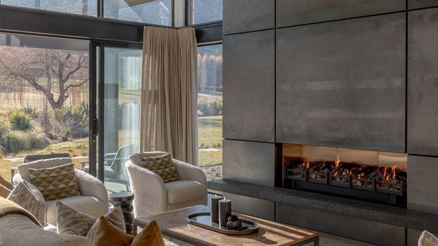 A refined contemporary living room with floor-to-ceiling glazing overlooking a landscaped garden. Two upholstered armchairs and a sofa surround a low timber coffee table, while a long linear fireplace is integrated into a dark tiled or concrete-look feature wall. Full-height curtains, natural light, and warm firelight create a sophisticated indoor-outdoor atmosphere.