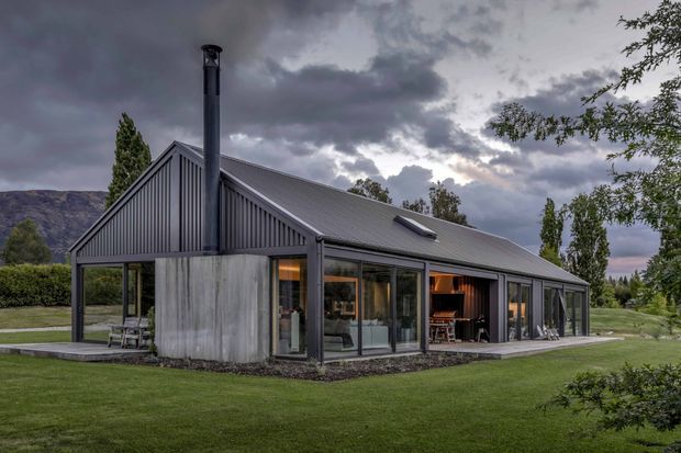 Exterior view of a contemporary single-storey gable-roof house set in a landscaped lawn, with dark vertical metal cladding, extensive floor-to-ceiling glazing, a long pitched roof, and a prominent metal flue. Warm interior lighting is visible through the windows and open glazed doors. Mountains and dramatic clouds form the distant background.