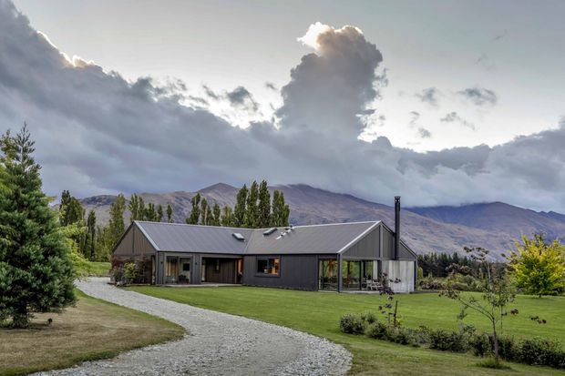 A single-storey contemporary rural home with dark vertical cladding and a low-pitched gabled metal roof, set within a landscaped lawn and gravel driveway. Large glazed openings connect the house to the garden, with dramatic mountains and mature trees forming the backdrop.