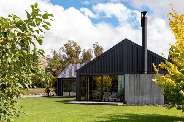 Exterior view of a contemporary single-storey house set within a landscaped lawn and trees. The home features dark vertically ribbed metal cladding, steep gabled roof forms, extensive dark-framed glazing, a large raw concrete wall, and a prominent black flue or chimney. A covered outdoor seating area is visible behind the glazing.