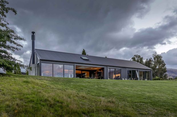 A low, elongated contemporary house set in a broad grassy landscape beneath dramatic clouds. The building has a dark pitched metal roof, extensive floor-to-ceiling glazing, a prominent chimney, and warm interior lighting visible through the open central section. Trees and distant hills surround the property.