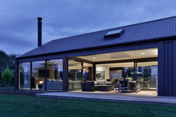 Dusk exterior view of a contemporary single-storey home with a dark standing-seam metal pitched roof, extensive floor-to-ceiling glazing, and a broad covered outdoor deck. The open living area, dining area, and kitchen are visible through the glazing, with warm interior lighting contrasting against the blue evening light. A dark vertical-clad wall and roof-mounted skylight are prominent architectural features.