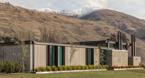 Single-storey contemporary residential building set against a rugged mountain landscape. The house has a low, elongated flat-roof form, vertical light-toned cladding, dark window and roof trims, large glazed openings, and a prominent dark masonry chimney or fireplace volume. A manicured lawn and planted border sit in the foreground, while snow-capped mountains dominate the background.