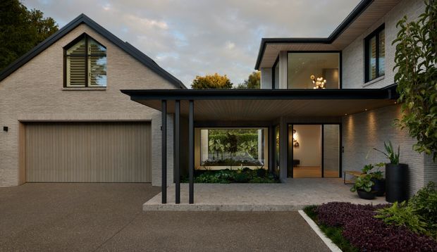 Contemporary two-storey residential home with a prominent gabled garage wing and a flat-roofed entrance addition. The facade combines pale textured masonry, dark charcoal rooflines and structural posts, large glazed openings, and a broad covered entry canopy. A double garage, landscaped garden beds, paved driveway, and warm interior lighting are visible.