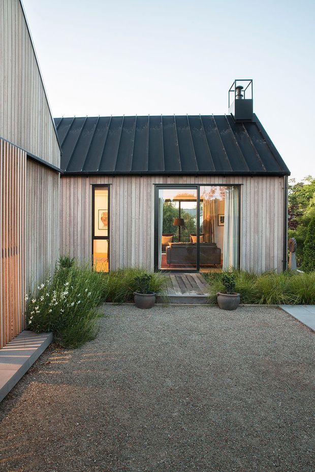 A compact modern dwelling viewed from a landscaped gravel courtyard. The building has vertically clad timber walls, a dark standing-seam metal pitched roof with a simple chimney enclosure, and large glazed doors and windows opening toward a small timber deck. Grasses and potted plants soften the gravel forecourt, while a warm, furnished interior is visible through the glazing.
