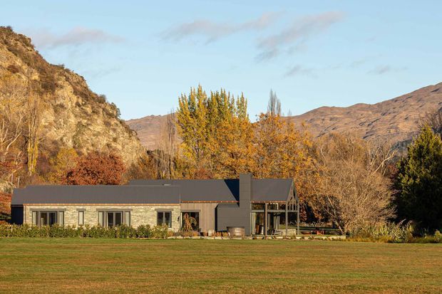 A single-storey contemporary home set against a rugged mountain landscape. The long, low facade combines pale masonry walls with dark pitched roofing, extensive glazing, and a prominent dark chimney or vertical feature. Autumn trees surround the house, with a broad lawn in the foreground and steep hills rising behind.