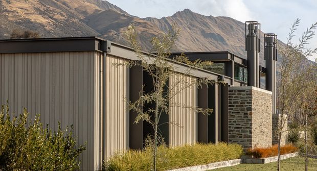 Exterior view of a contemporary single-storey building set against steep mountain terrain. The facade combines vertically ribbed taupe cladding, dark metal roof and trim, extensive glazing, and a prominent rough stacked-stone wall with tall dark metal chimney elements. Native-style grasses and sparse landscaping frame the building.
