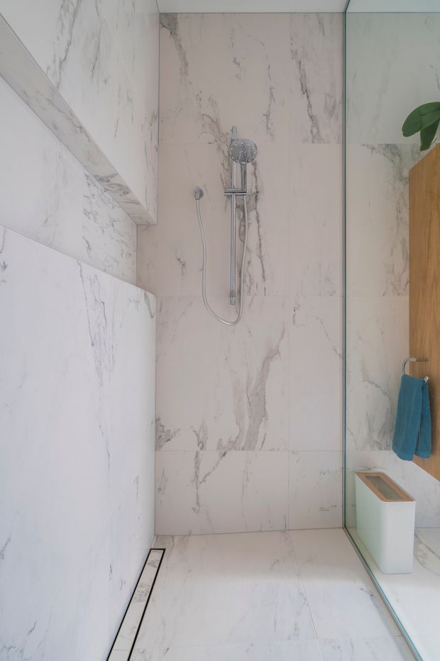 A minimalist walk-in shower bathroom clad almost entirely in white marble-look slabs with grey veining. The frameless glass shower screen, wall-mounted handheld shower, linear floor drain, and light timber vanity edge are visible, with a small turquoise towel adding colour.