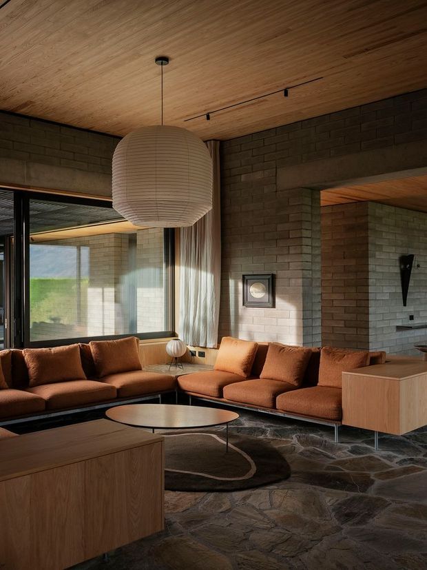 Warm, moody contemporary living room with a large rust-brown sectional sofa, low round coffee table, timber built-in cabinetry, and a sculptural paper pendant light. The room features exposed dark brick masonry walls, a timber-lined ceiling, expansive glazing with curtains, and an irregular natural stone floor.