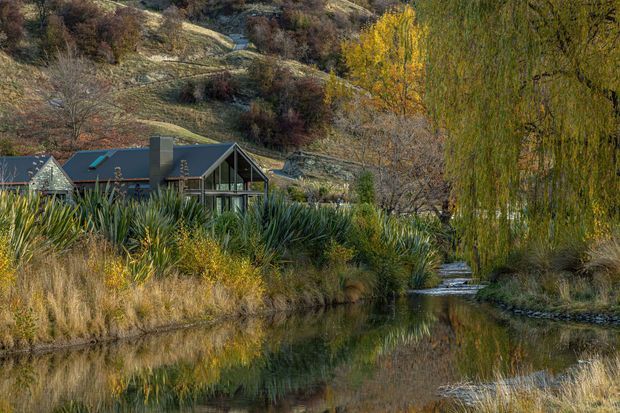 A contemporary gabled house set within a natural, landscaped rural or bush setting beside a reflective pond or slow-moving stream. The dark-roofed building has extensive glazing and a prominent triangular gable, while native-looking grasses and sculptural plantings frame the water. Autumn foliage and a steep rocky hillside form the background.