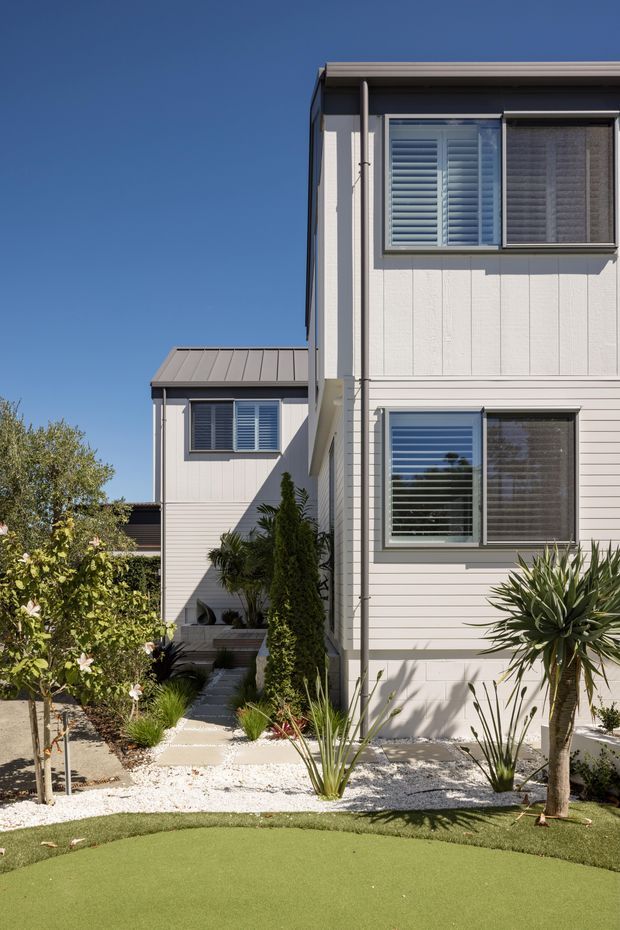 Exterior view of a contemporary multi-storey residential building with light-coloured horizontal cladding, vertical panel sections, metal roofing, and multiple windows fitted with external louvred shutters. A landscaped front garden includes white gravel, paved stepping paths, sculptural drought-tolerant plants, and a manicured artificial-turf area in the foreground.