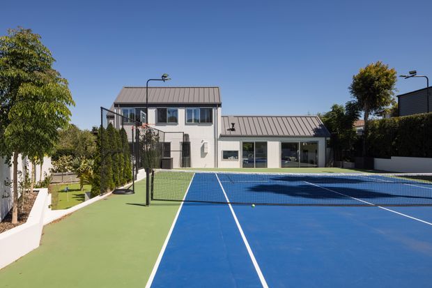 A bright exterior view of a modern multi-level residential property with a large blue and green tennis court in the foreground. The building has simple white rendered walls, dark metal pitched roofs, extensive glazing, and contemporary rectangular forms, with landscaped planting and boundary hedges around the court.