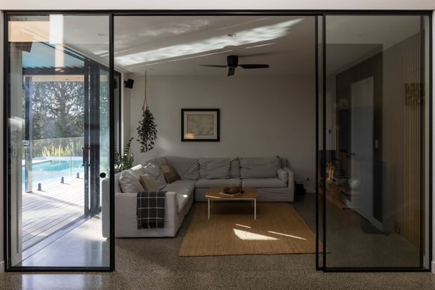 A contemporary living room viewed through large black-framed glazed doors. The room contains a grey sectional sofa, matching armchair, low timber coffee table, natural woven rug, ceiling fan, and minimal wall art. A glazed opening leads to a timber deck and swimming pool outside, while the dark-framed glazing creates strong reflections and a clear indoor-outdoor connection. The palette is neutral and warm, with natural textures and abundant daylight.