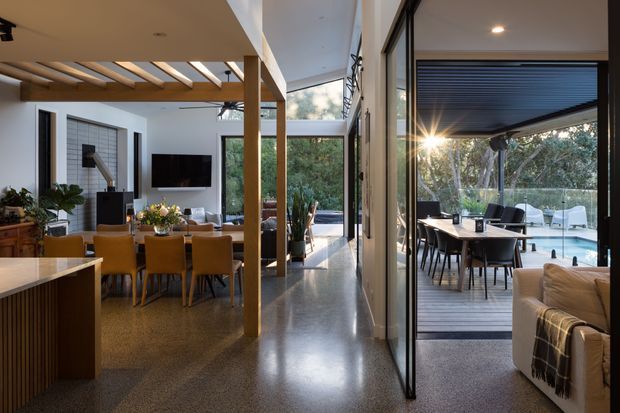 A contemporary open-plan residential interior with a dining area, living room and kitchen arranged around a glazed courtyard or outdoor entertaining space. Timber posts and a slatted pergola structure frame the space, while extensive floor-to-ceiling glazing connects the interior to an outdoor dining area and swimming pool. The interior features polished concrete or terrazzo-like flooring, white walls, warm timber joinery, and strong indoor-outdoor connection.
