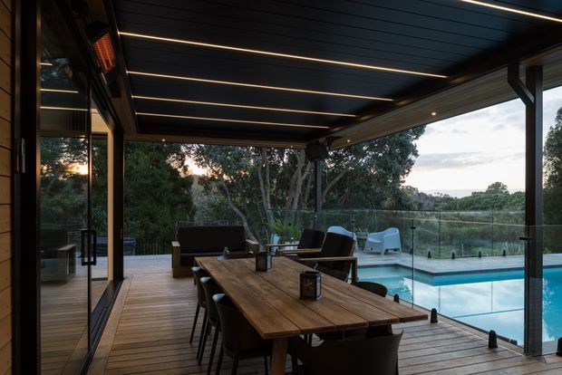 Covered outdoor entertaining area with a timber dining table and seating on a timber deck beside a swimming pool. The space has a dark linear-panel ceiling with integrated strip lighting, slender dark structural posts, extensive glazing, and views to a leafy landscape. A barbecue or outdoor heating element is visible along the side wall, creating a refined indoor-outdoor entertaining setting.