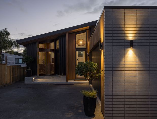 A dusk photograph of a contemporary residential facade and entry courtyard. The home features dark vertical cladding, warm timber detailing, screened entry doors, tall glazing, and a projecting grey masonry-tile wall with integrated up-and-down exterior lighting. Concrete paving, fencing, and potted plants frame the approach.