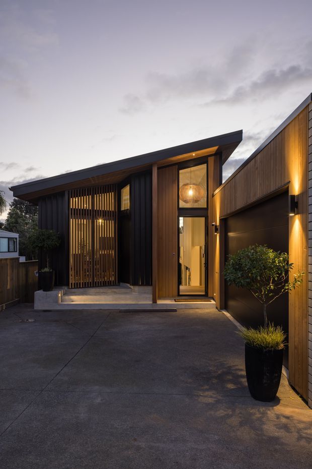 Dusk exterior view of a contemporary residential entrance and attached garage. The facade combines dark vertical cladding, warm timber battens and wall panels, extensive glazing, concealed warm lighting, and a sharply projecting low-pitched roof. A paved driveway leads to the recessed front door and garage, with simple potted planting at the edges.