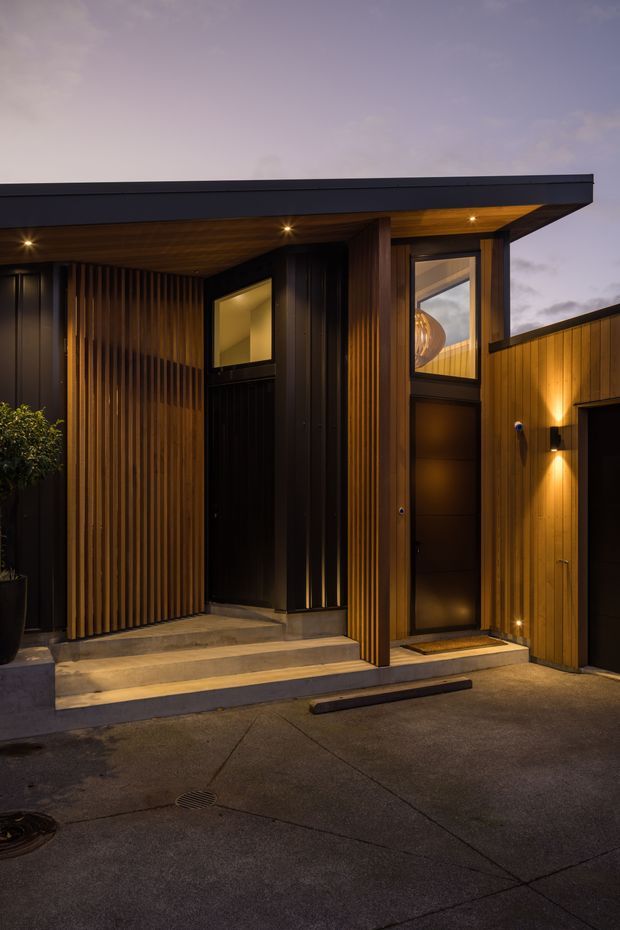 Twilight exterior view of a compact contemporary residential entry and attached garage. The facade combines dark vertical timber slats and dark metal cladding beneath a broad low-pitched flat roof, with warm recessed and wall-mounted lighting, concrete entry steps, and a dark timber front door.
