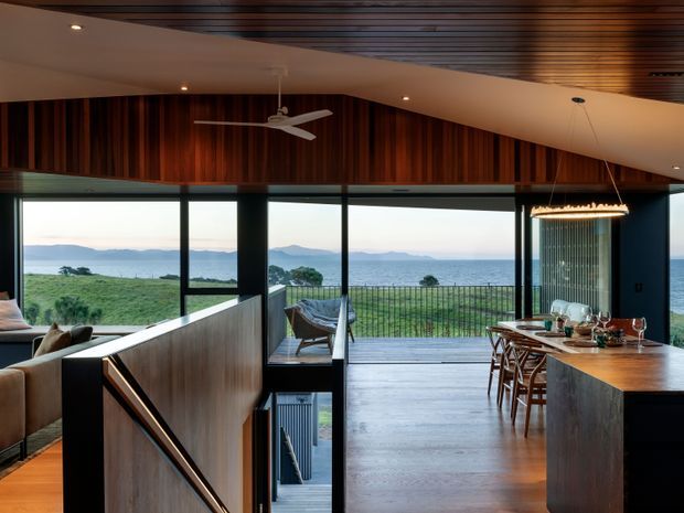 Open-plan coastal interior combining a dining area and kitchen with a visible stairway and adjacent living space. Full-height glazing frames ocean and coastal landscape views, while dark timber wall panelling, a slatted timber ceiling, warm timber flooring, and a sculptural pendant create a refined, warm atmosphere.
