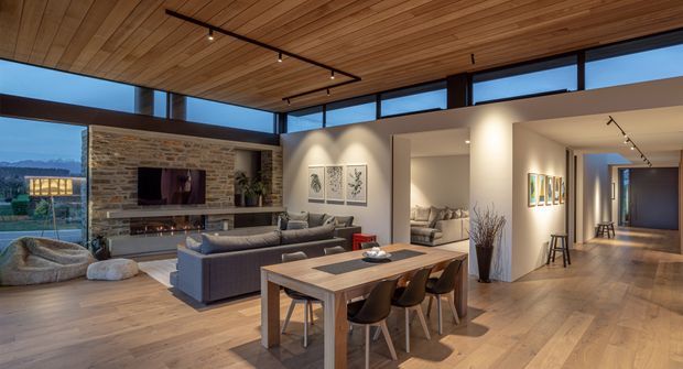 Open-plan contemporary living and dining area with a warm timber-lined ceiling, extensive clerestory glazing, wide timber flooring, a stone feature fireplace wall, grey sofas, and a timber dining table. The room opens toward a hallway and appears to overlook a landscaped setting and distant hills.