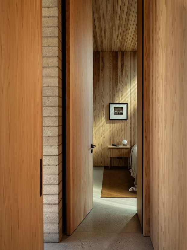 A narrow interior passage framed by tall natural timber doors and wall panels, leading into a warm timber-lined bedroom. The bedroom contains a partially visible bed, a small timber desk or console, framed artwork, a rug, and soft indirect daylight. A vertical strip of light-coloured masonry blocks contrasts with the extensive wood surfaces, while carpet continues through the hallway.